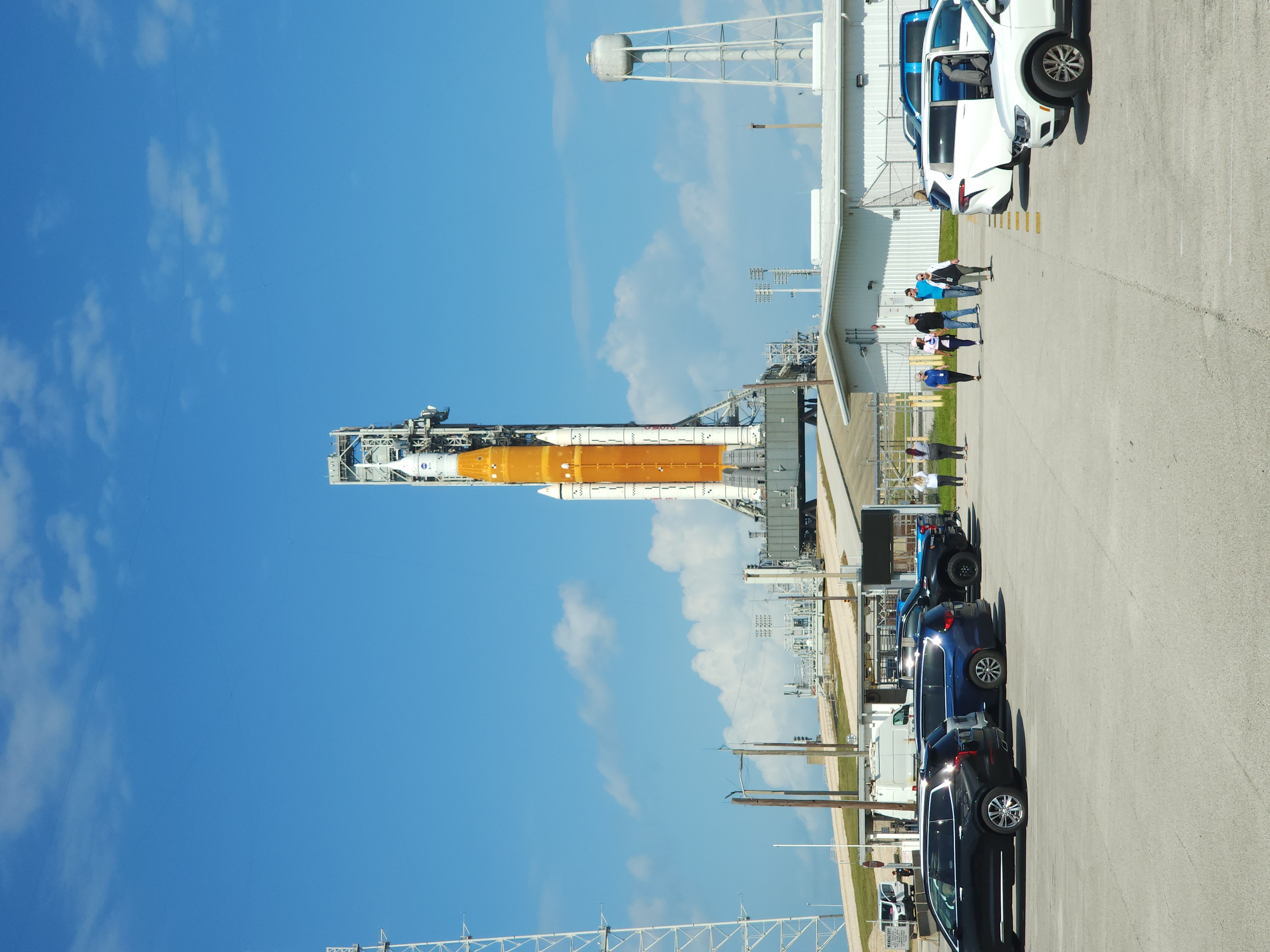 SLS rocket zoomed out view from the parking lot in front of a blue sky.