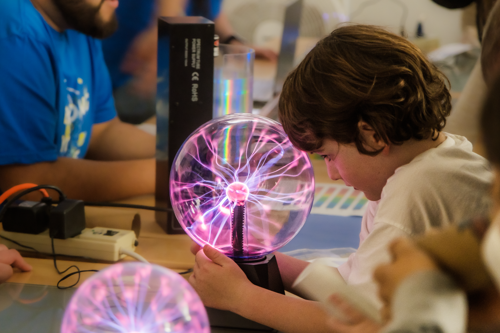 A child places their forehead on a Tesla Ball and grasps its sides, looking into the inside and watching the plasma at work.